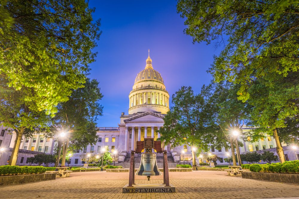 Bell sits in front of an illuminated domed building surrounded by trees at dusk. Text: "WEST VIRGINIA".