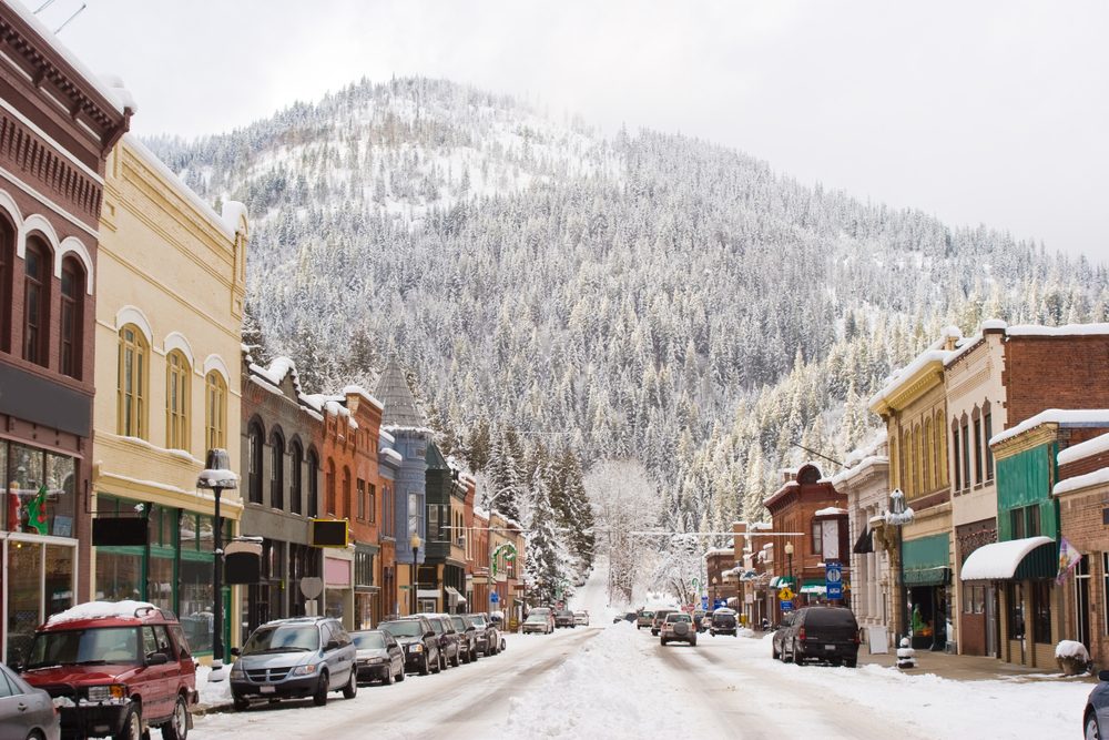 Buildings line a snow-covered street bustling with cars, framed by a forested mountain in the background on a winter day.