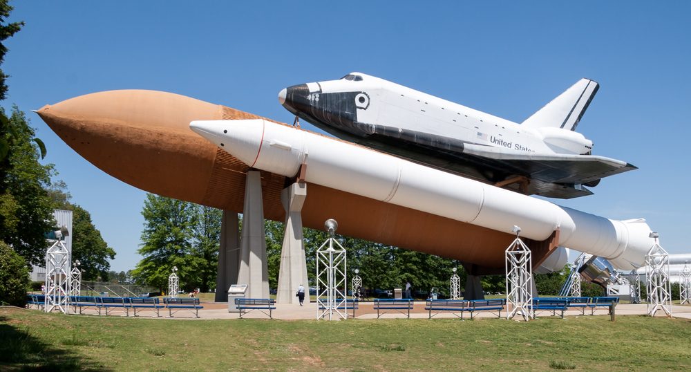 Space shuttle displayed on large boosters, elevated on stilts in a park-like setting with trees, benches, and a clear blue sky.