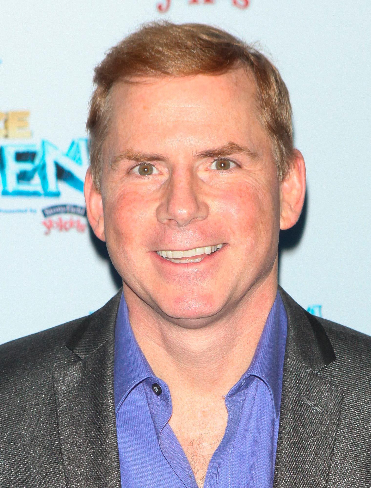 A person smiles directly at the camera, wearing a purple shirt and suit jacket against a light background with partially visible text.