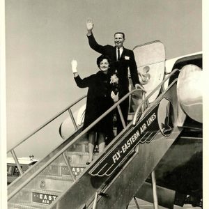 Two people wave at the top of airplane stairs with 