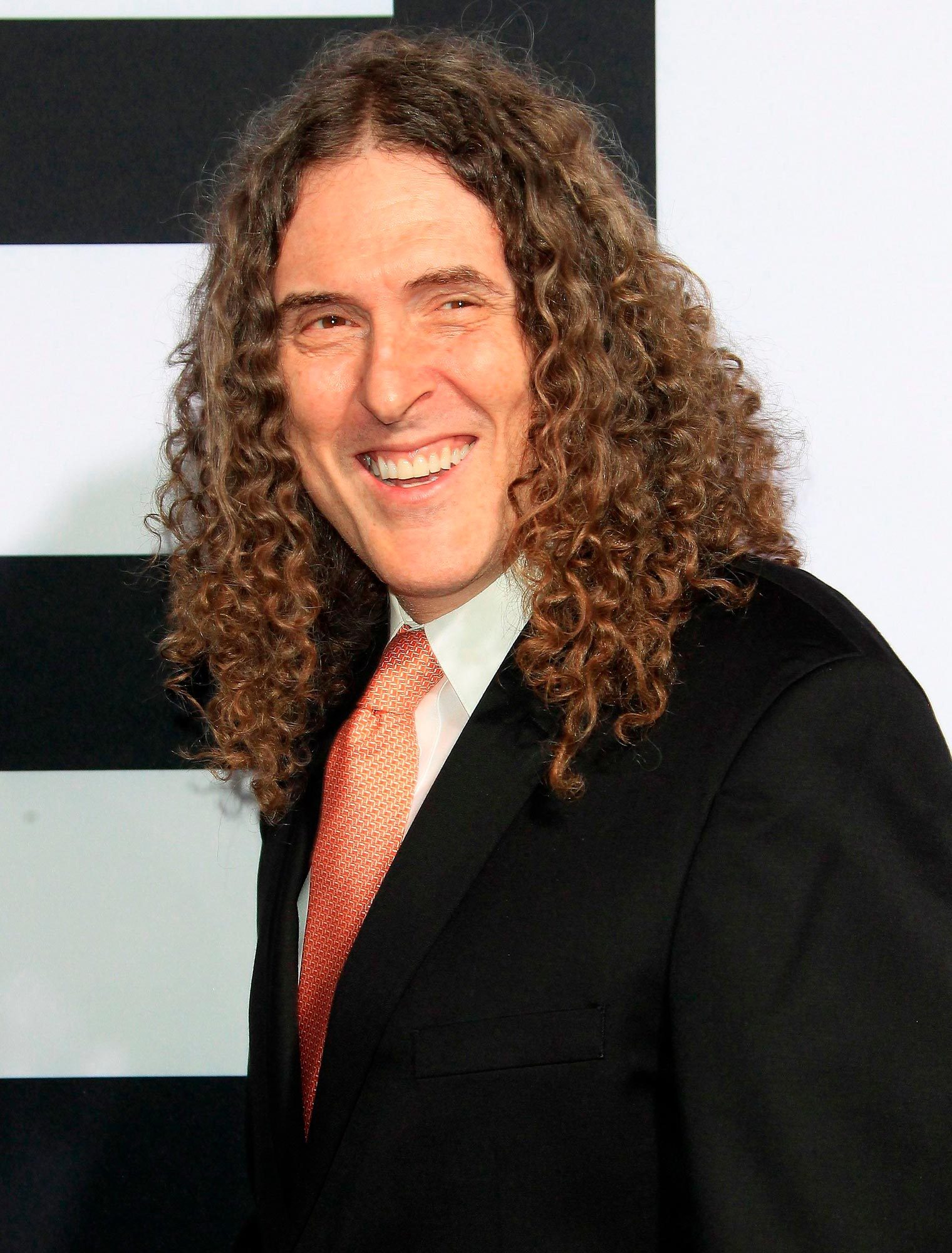 A person with long curly hair smiles, wearing a suit and orange tie, standing against a striped background.