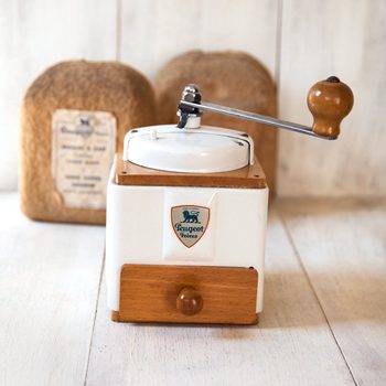 A vintage coffee grinder with a wooden handle stands on a wooden surface, with labeled coffee bags in the background. Text includes "Peugeot FrÃ¨res."