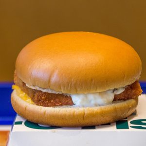 A burger sits on a box, featuring a fish patty and tartar sauce, against a plain, brown background.