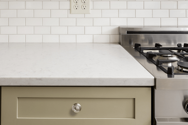 A white countertop beside a stainless steel stove, with a tiled backsplash and electrical outlet in a kitchen setting.