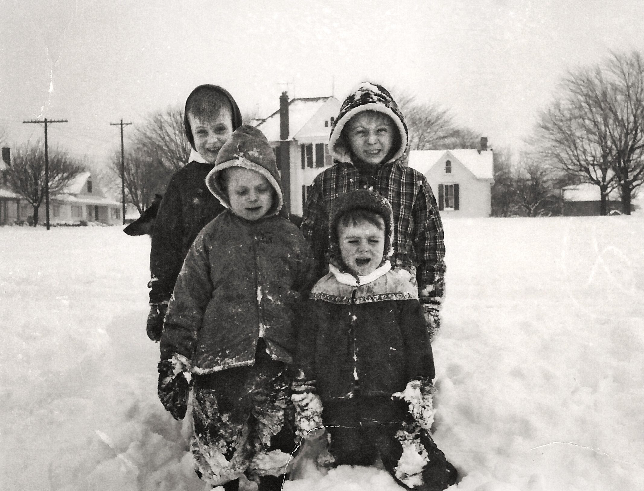 Four children in winter coats stand in deep snow, smiling, in front of snow-covered houses and leafless trees.