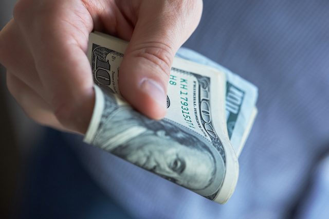 A hand holds multiple US $100 bills in a slight grip against a blurred blue-striped background.