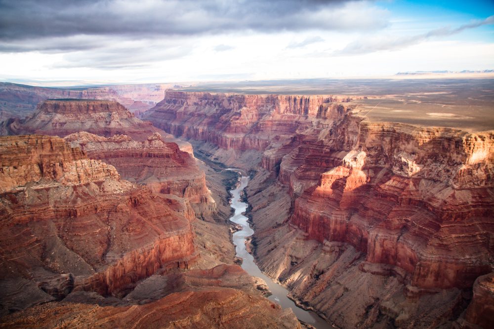 Canyon extends downwards, revealing layered rock formations, with a river weaving through the deep valley, set under a cloudy sky.