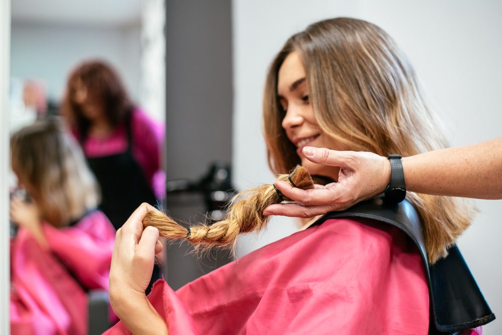 Hairdresser trims a smiling client's braid in a salon, with both wearing pink capes.