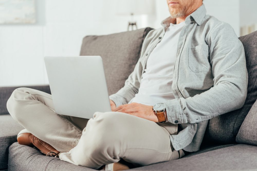 A person uses a laptop while sitting cross-legged on a gray sofa in a well-lit, modern living room.