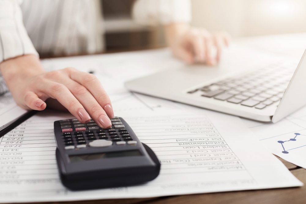Hand uses calculator on printed spreadsheets, next to an open laptop on a desk.