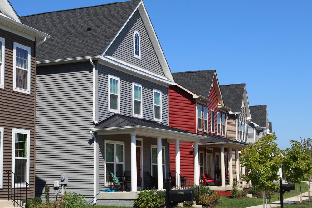 Houses line a street, featuring colorful facades, porches, and black mailboxes, with a backdrop of clear blue sky and greenery.