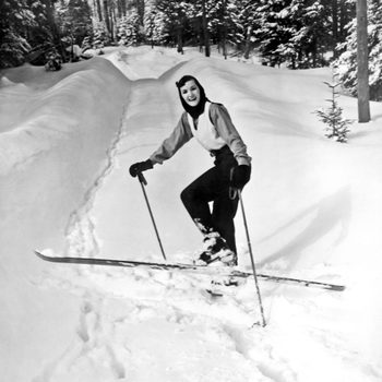 A person skis downhill on a snowy path surrounded by snow-covered trees, smiling and wearing ski gear.