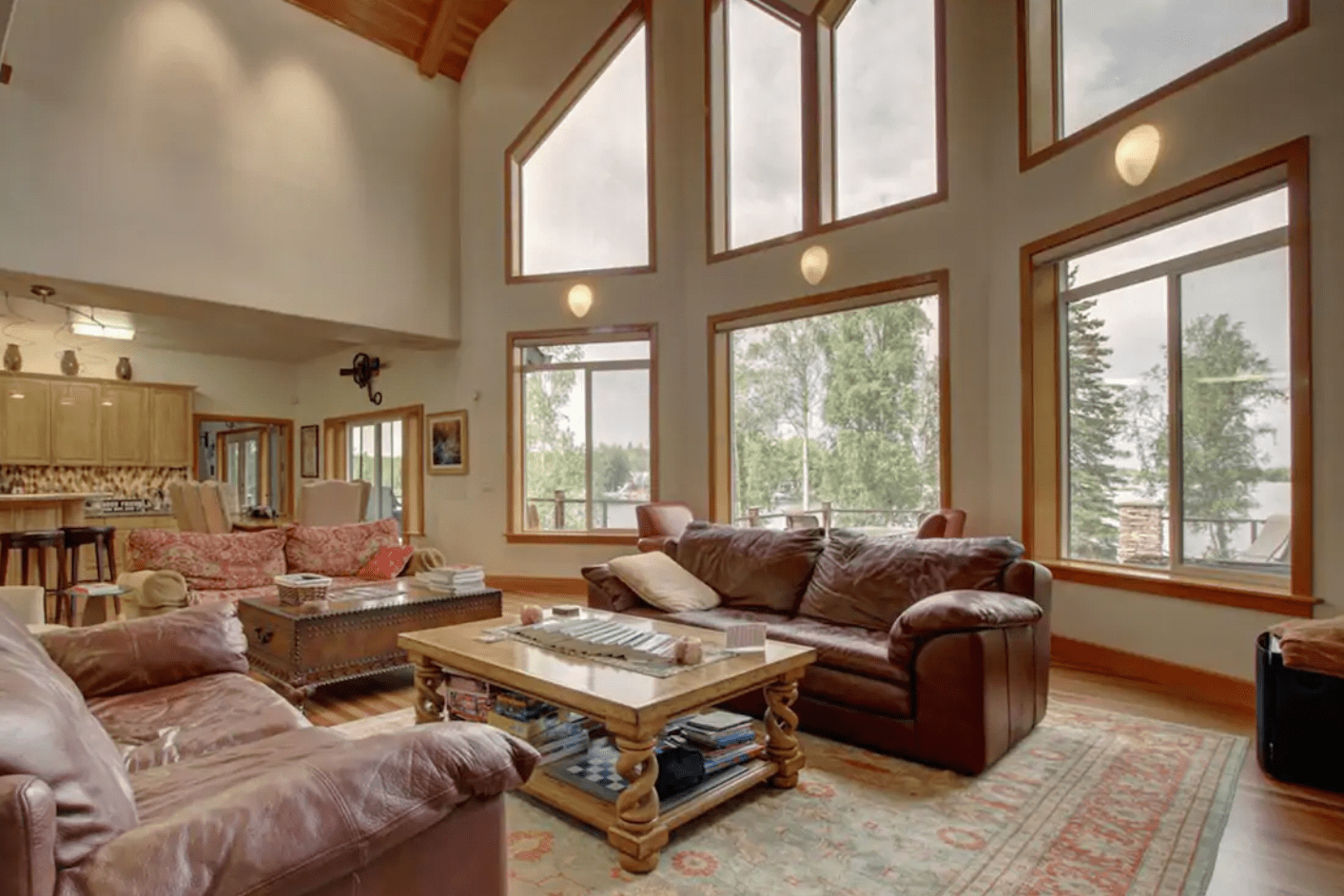 Sofas surround a wooden coffee table in a spacious living room with large windows and vaulted ceilings, overlooking greenery outside.