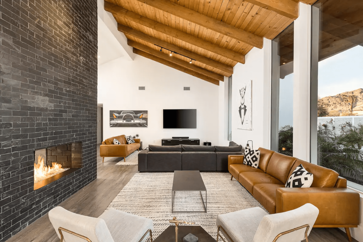 Fireplace glows warmly in a modern living room with brown sofas, a wall-mounted TV, large windows, and vaulted wooden ceiling, creating a cozy atmosphere.