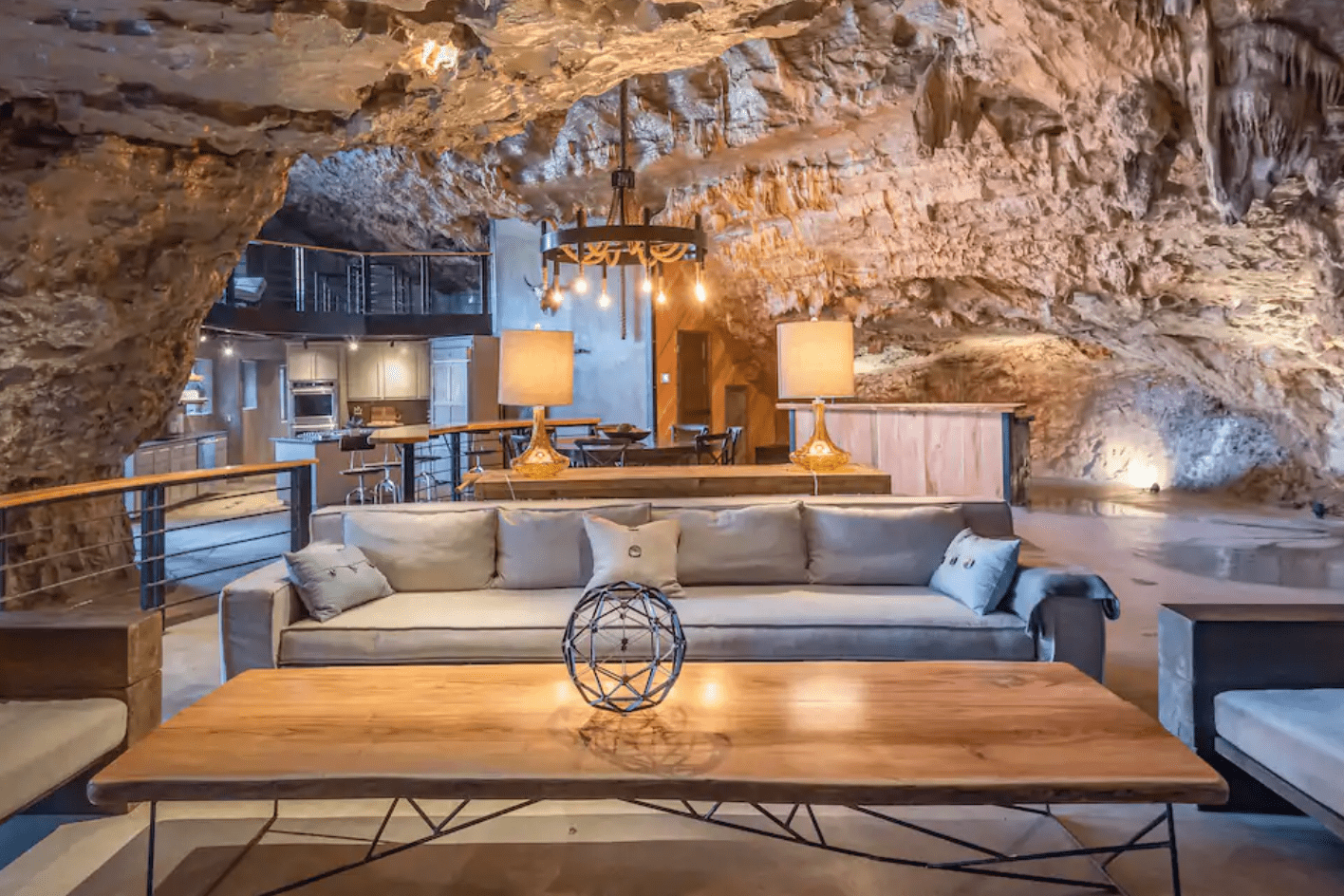 A modern living room is set inside a cave, featuring a wooden table and gray sofa, with warm lighting and rustic rock walls.