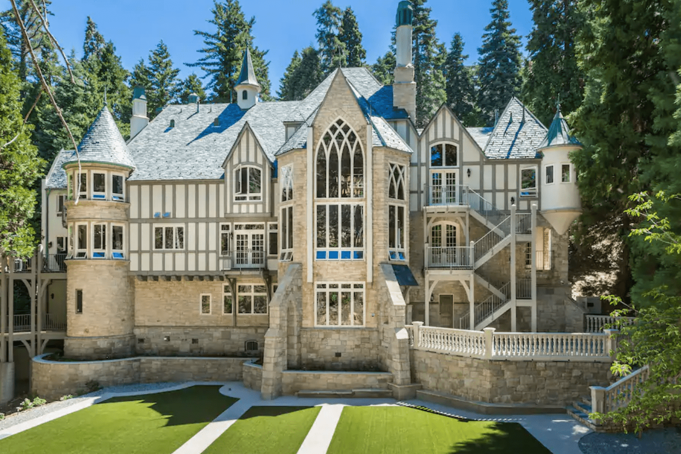 A large stone mansion displays gothic architecture with pointed spires and arched windows, surrounded by lush green trees and a manicured lawn.