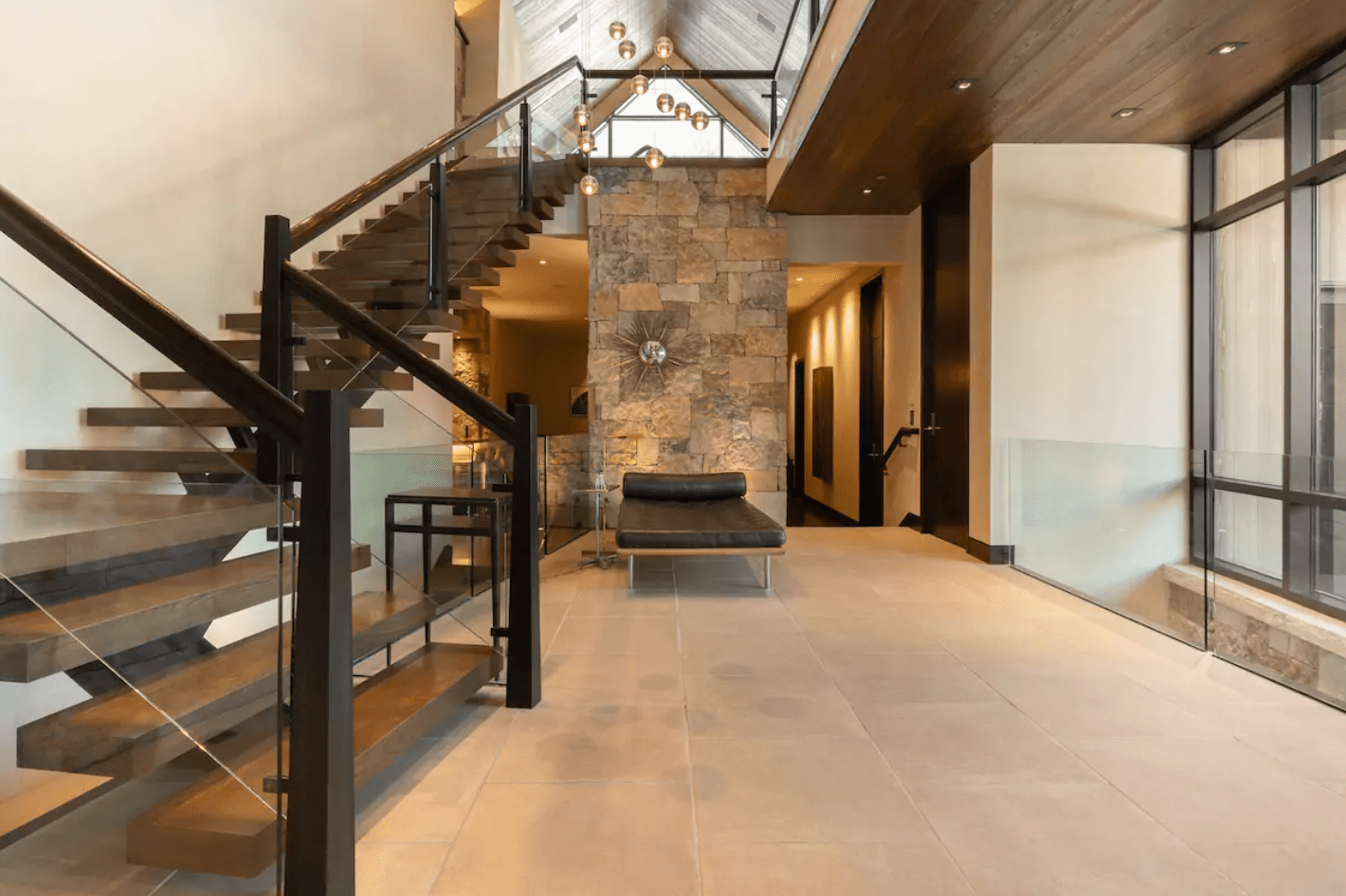 Floating wooden staircase ascends alongside a stone wall, with modern seating below in a spacious, contemporary hallway featuring glass and wood accents.
