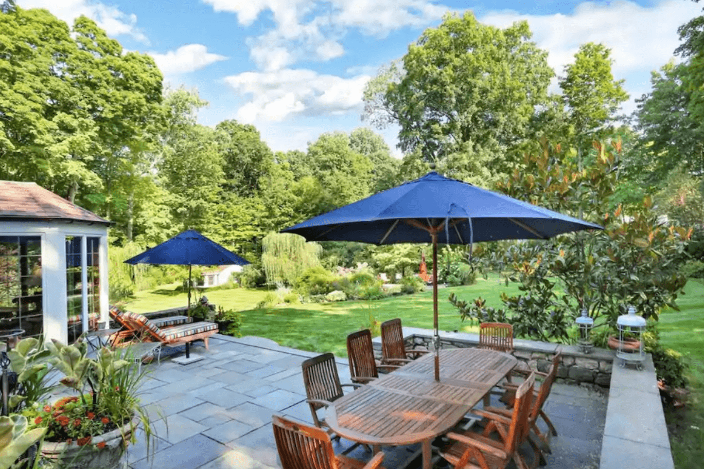Outdoor wooden table sits under blue umbrella on patio, surrounded by lush garden with trees under a clear sky.