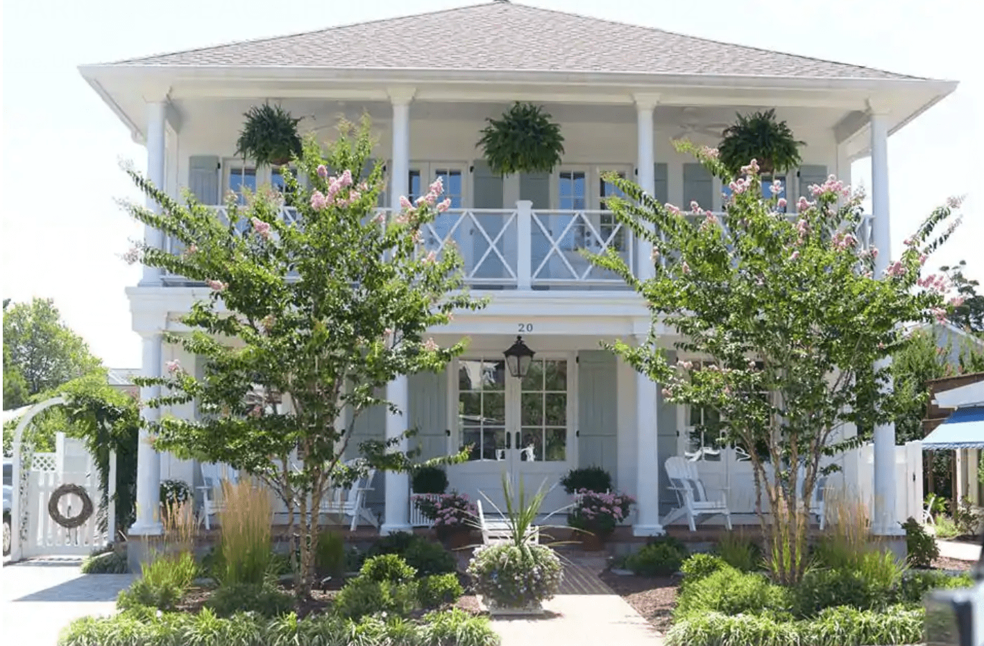A two-story house with wraparound porch features hanging plants and flowering trees, nestled in a landscaped garden. Number "20" is displayed near the door.