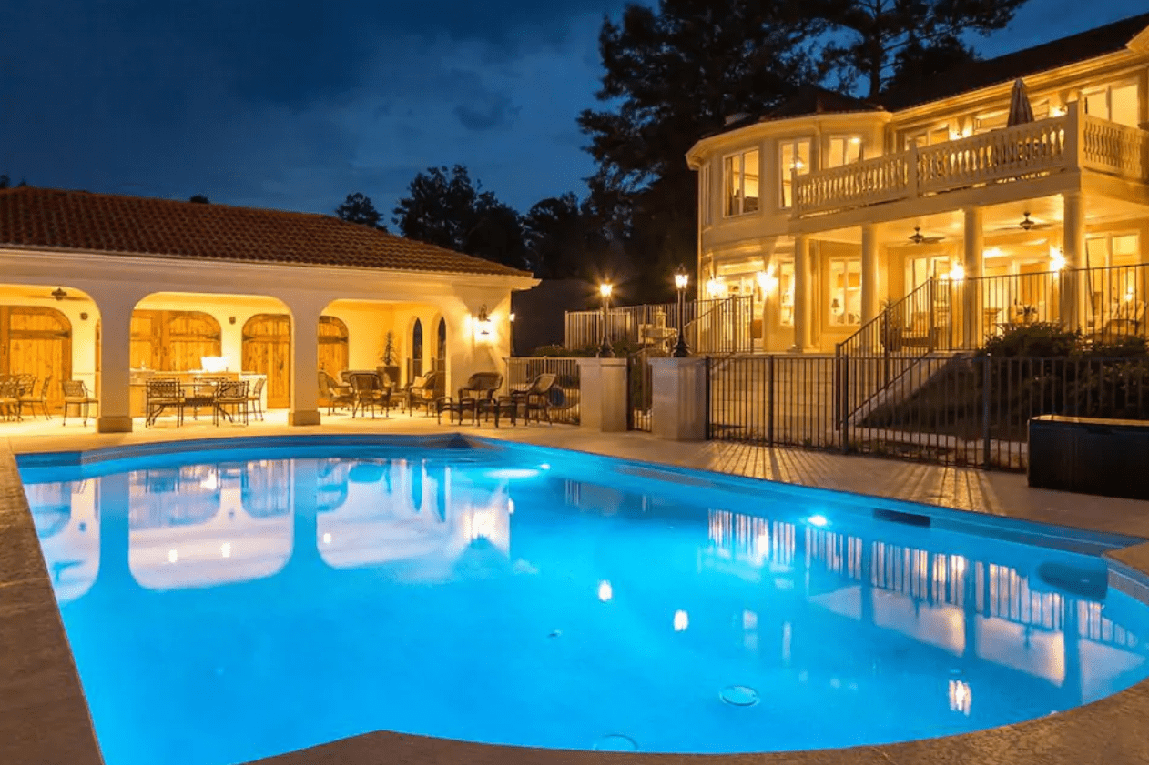 A brightly lit swimming pool reflects a luxurious mansion at night, with arched verandas and warm lights illuminating the surrounding area.