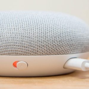 A gray smart speaker rests on a wooden surface with a visible orange mute button and charging cable connected at the back.