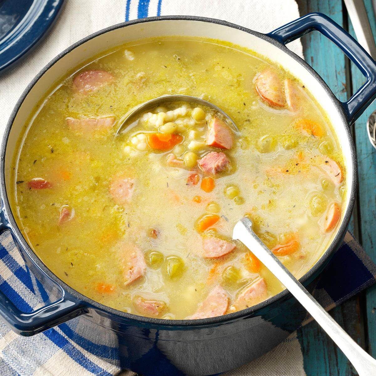 A pot contains thick, hearty soup with chunks of sausage, carrots, and peas, sitting on a blue striped cloth.