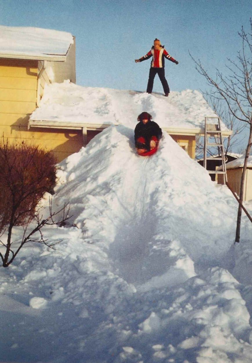 Person sleds down roof on snow pile; another stands above with arms outstretched. Snow-covered house and trees surround them.