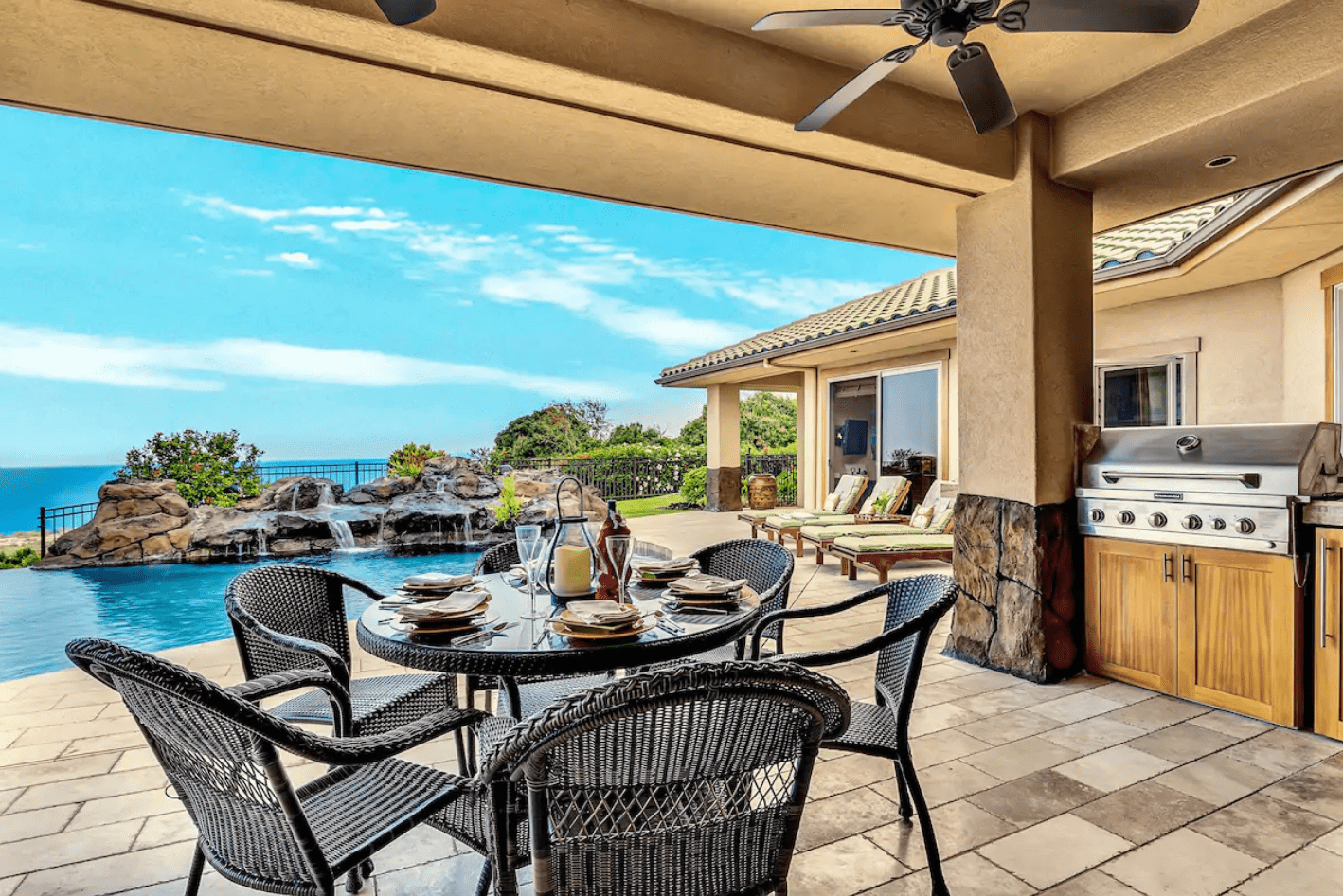 Patio table holds dishes near pool with waterfall; barbecue grill beside lounge chairs under sunny sky with ocean view.