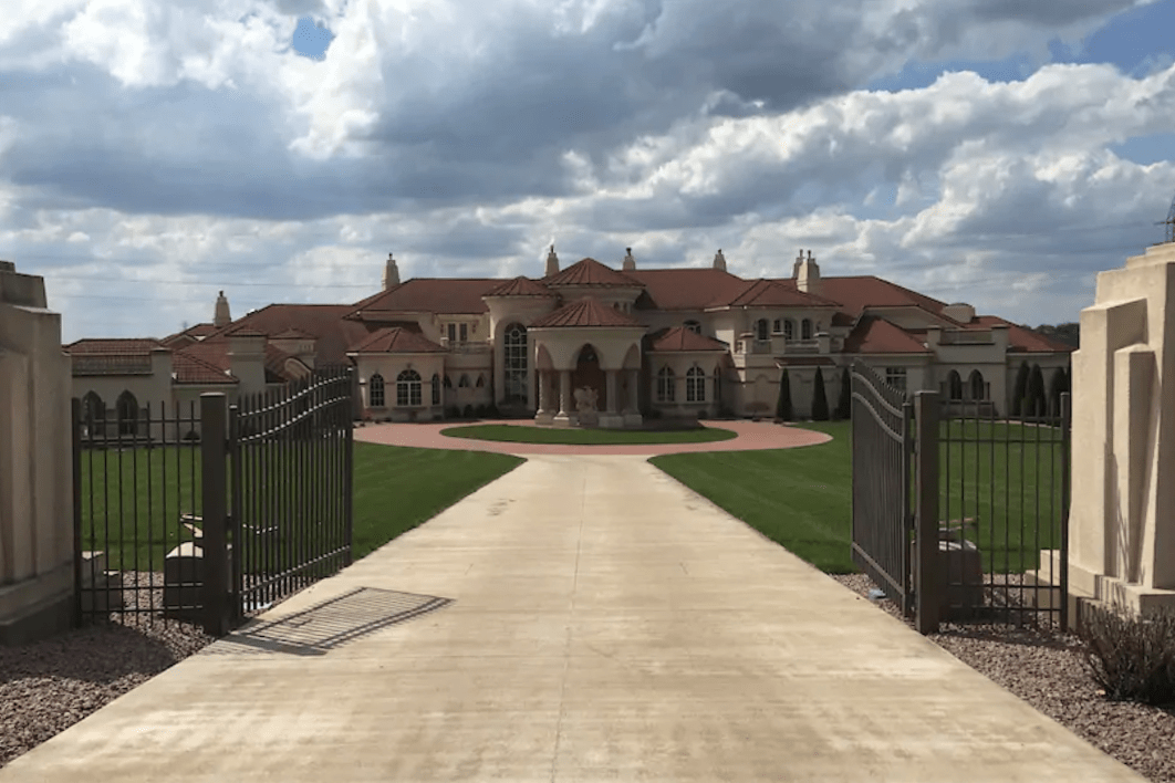 Mansion stands surrounded by open wrought-iron gates, on a wide driveway under a partly cloudy sky.