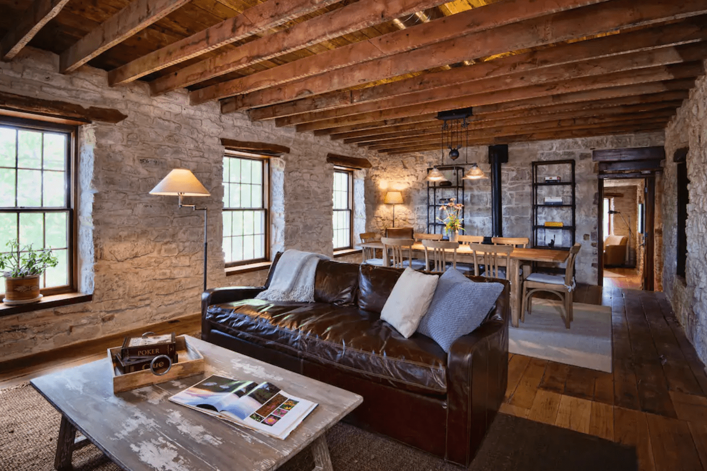 Leather couch sits in rustic living room; wooden beams, stone walls, and a dining table enhance cozy ambiance. Books and decor are placed nearby.