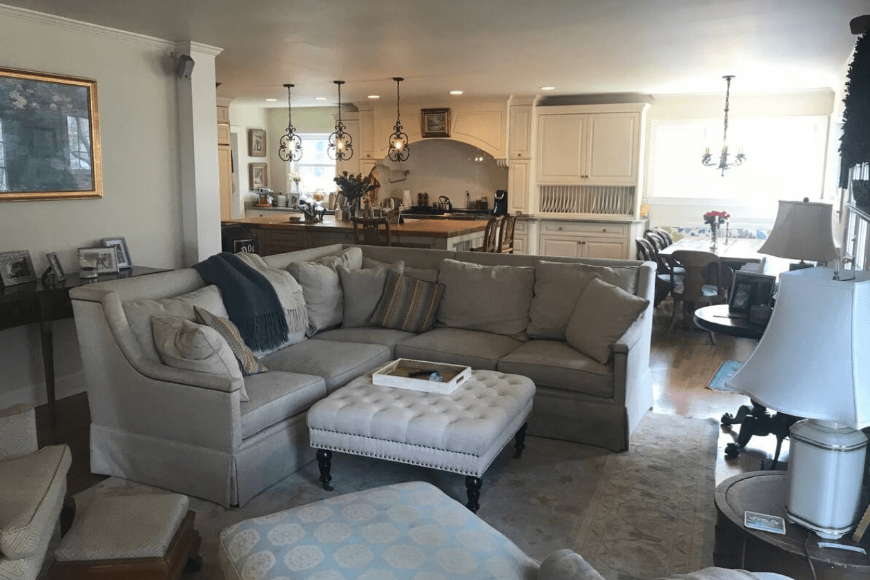 A beige sectional couch sits with pillows in a living room, adjacent to a kitchen, under decorative chandeliers and beside framed art.
