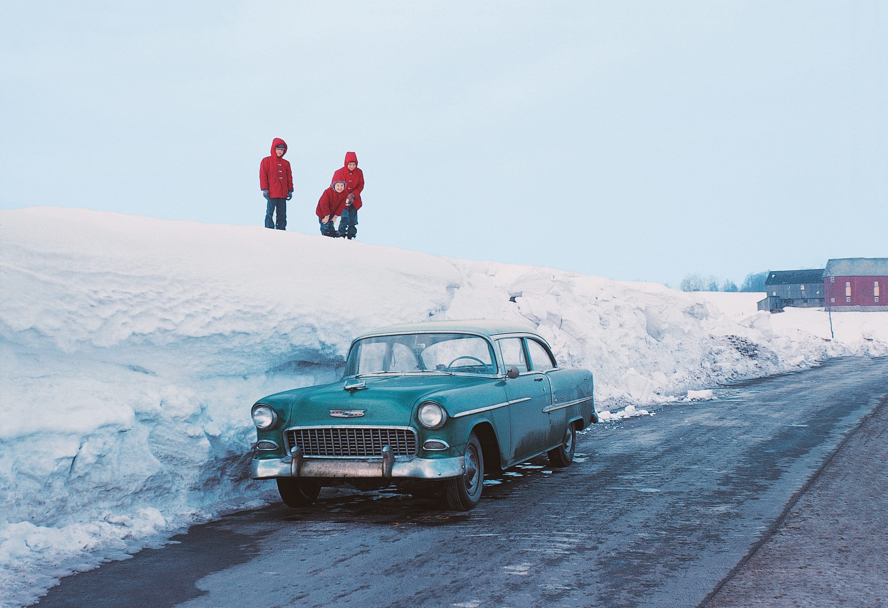 A turquoise vintage car parked beside snowbanks; three children in red coats stand atop the snow. Barns visible in the background.
