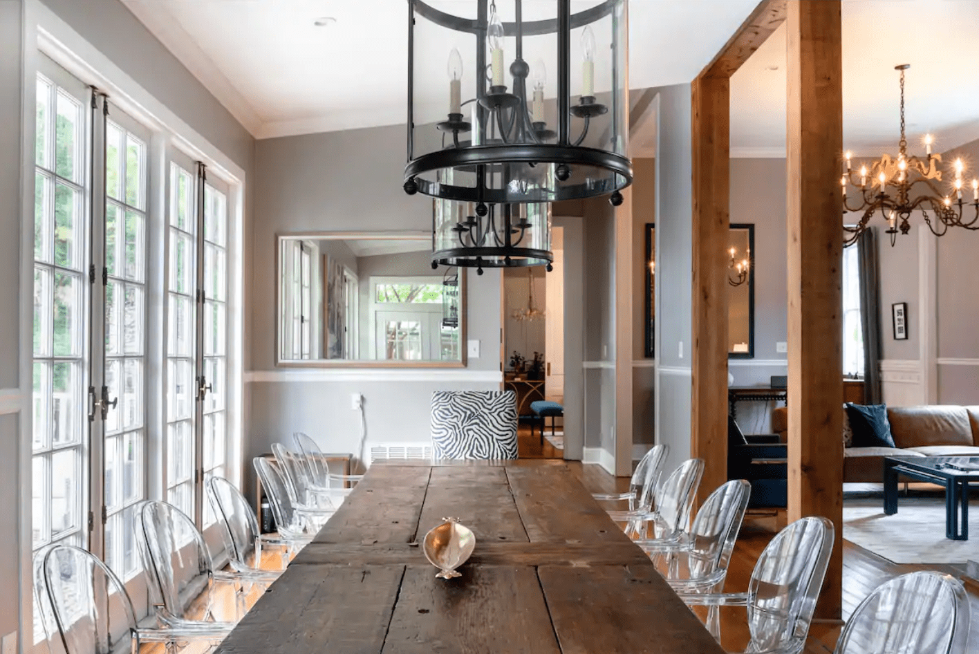 A long wooden dining table with clear chairs sits beneath modern chandeliers in a bright, spacious room with large windows and wooden beams.