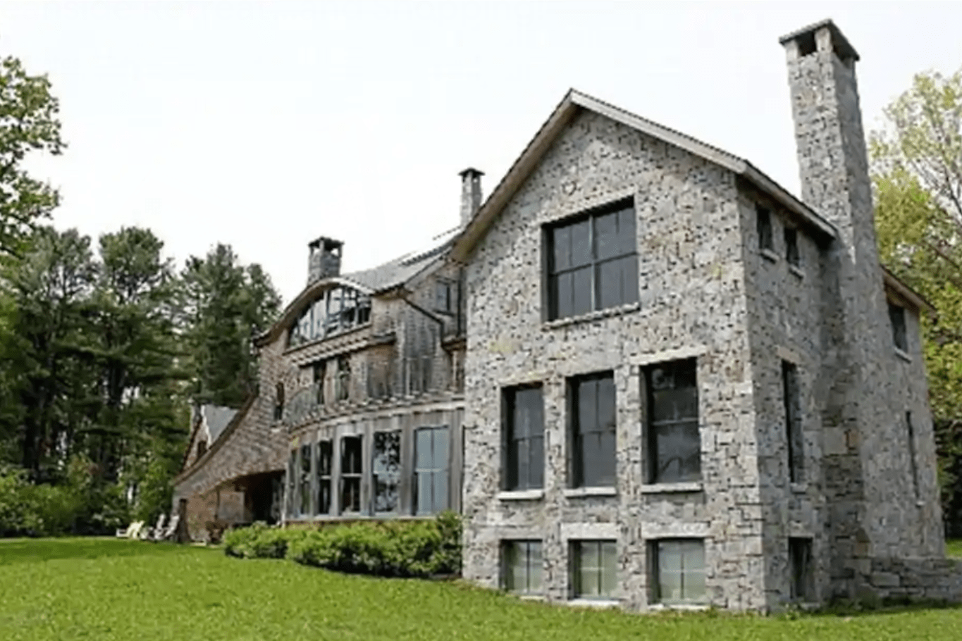Stone house stands with large windows and chimneys, surrounded by green lawn and trees under a clear sky.