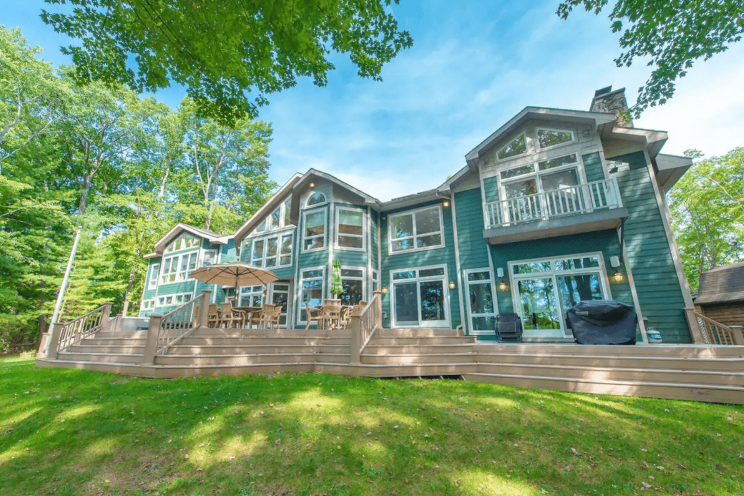 Large green house, surrounded by trees, features expansive deck with tables, chairs, and an umbrella under a clear blue sky.