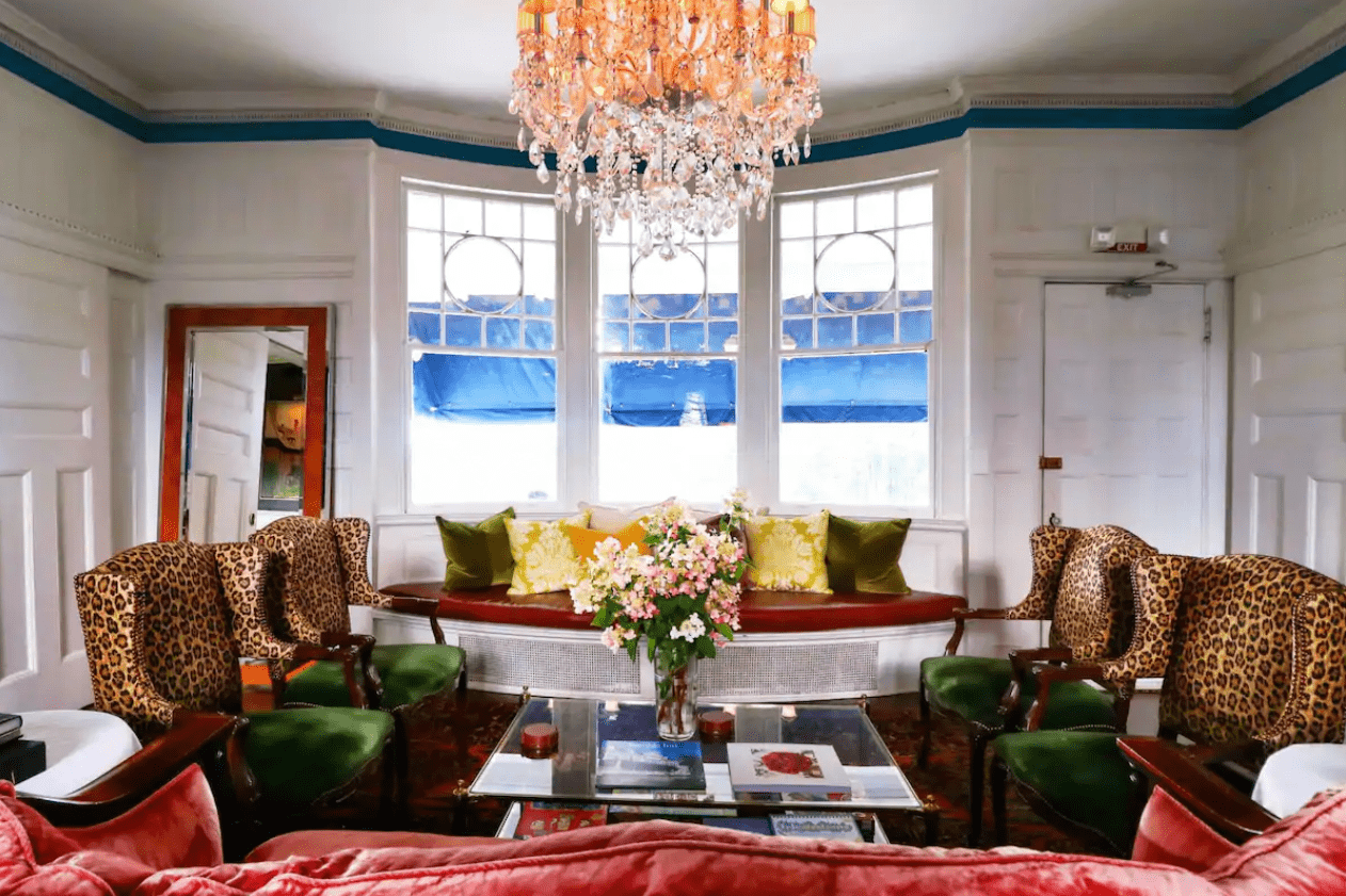 Chandelier hangs in an elegant room, featuring leopard-print chairs, green cushions, a glass coffee table with flowers, and large windows with blue awnings.