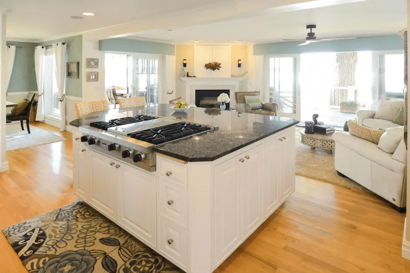 Kitchen island with stove; spacious open-plan room; hardwood floor; adjacent sitting area with white sofas; large windows; modern decor; cozy, light-filled atmosphere.