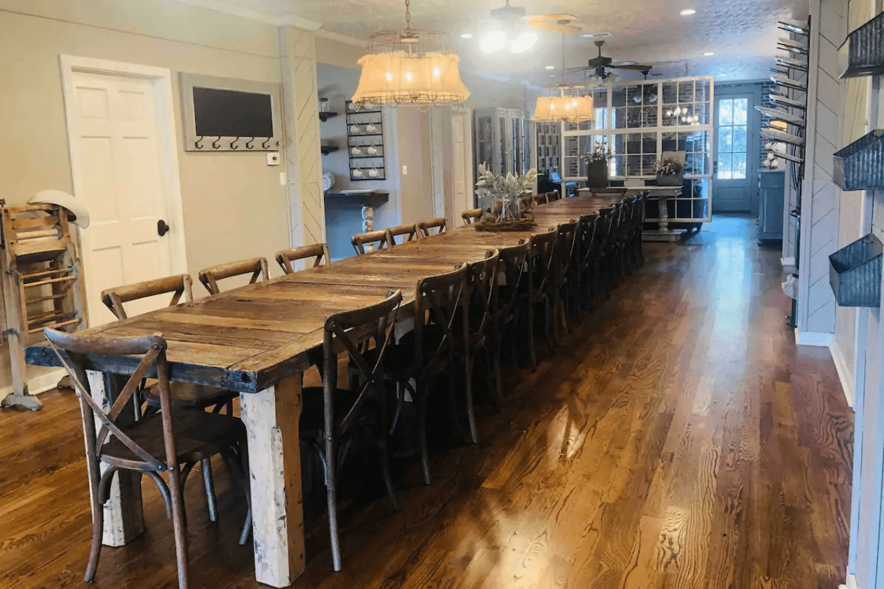 A long wooden table, surrounded by chairs, stretches along a wooden-floored dining room with pendant lighting and a shelving unit in the background.