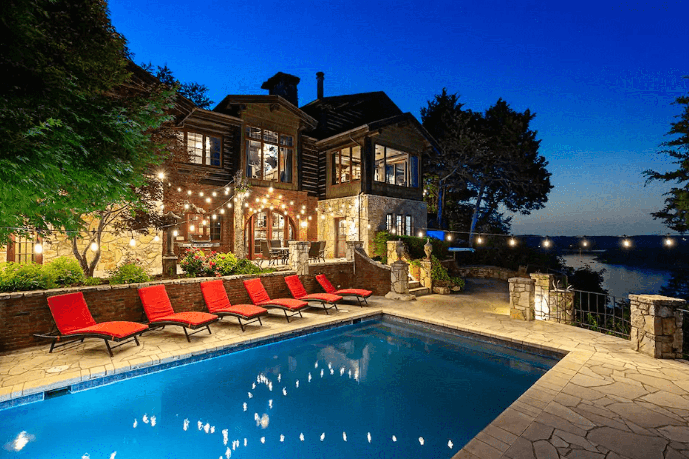 Pool reflects string lights; surrounded by red lounge chairs. Stone house with large windows and balcony overlooks it. Trees and lake in the background.