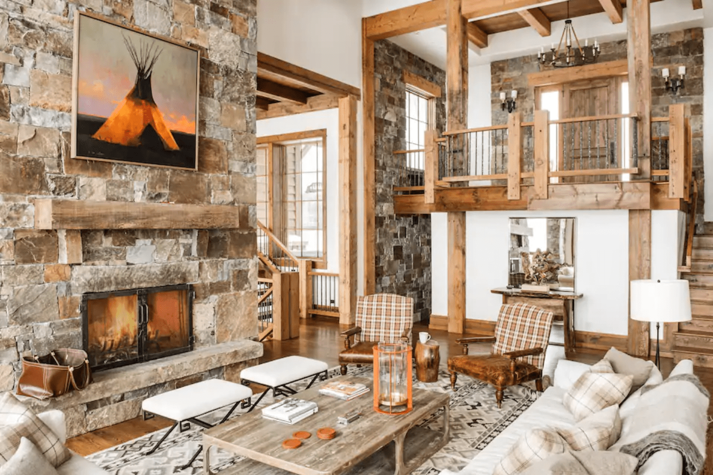 A cozy living room features a stone fireplace, wood beams, plaid chairs, and rustic decor, creating a warm, inviting atmosphere. A painting hangs above the fireplace.