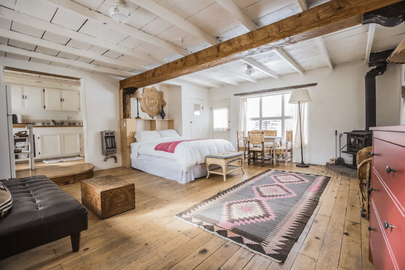 Bedroom suite with a bed, red chest, wood stove, and table, situated in a rustic, white-walled, wooden-floored open space.