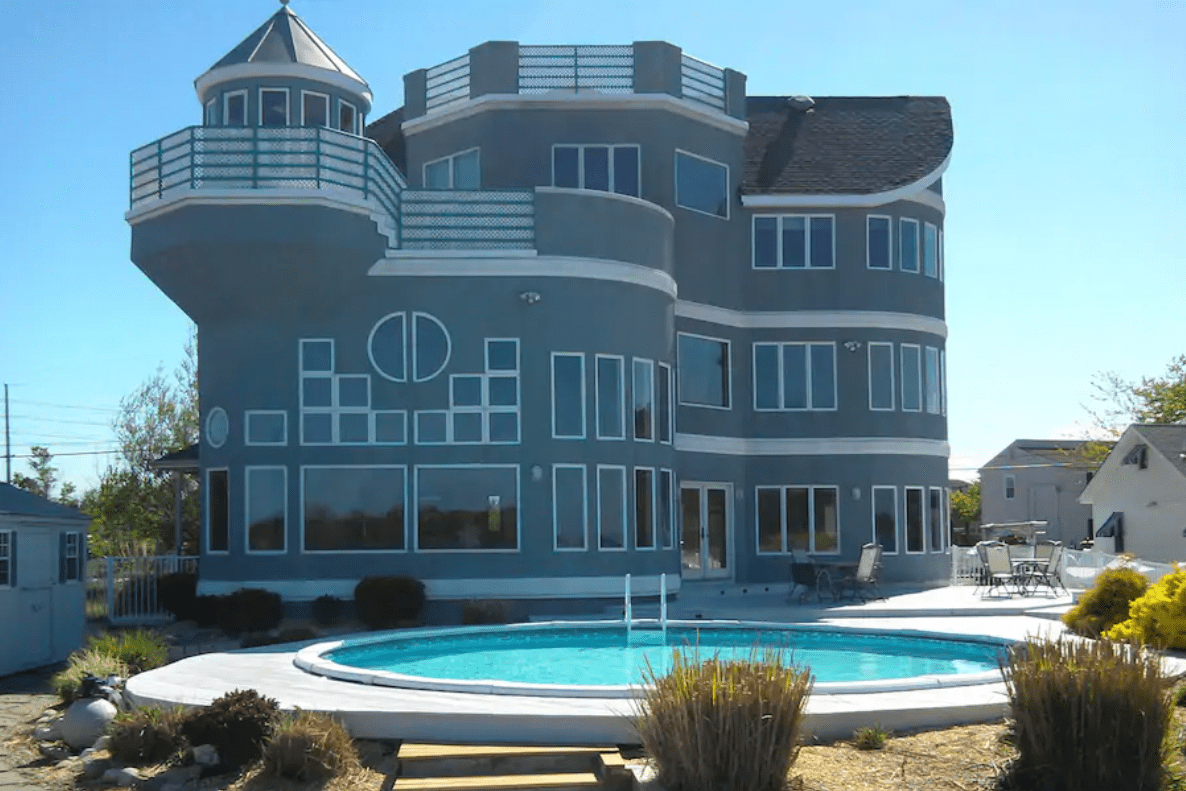A unique, multi-story blue house overlooks a round swimming pool, surrounded by a patio with outdoor furniture and landscaping in a residential area.
