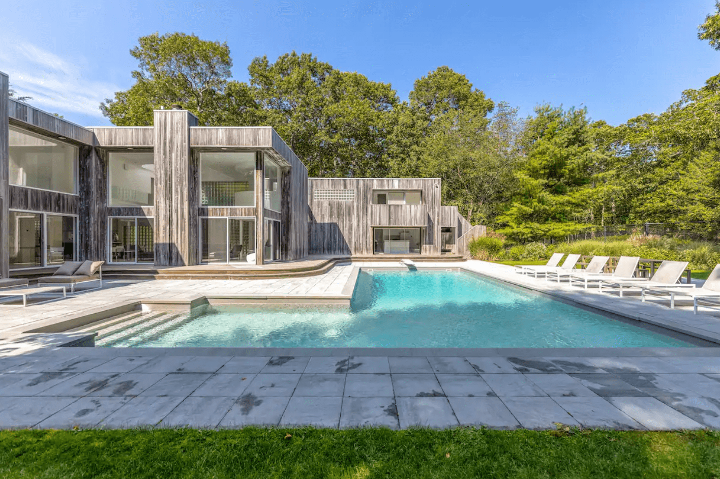 Modern house stands with wooden facade, overlooking a clear swimming pool surrounded by lounge chairs, set against a backdrop of lush green trees.