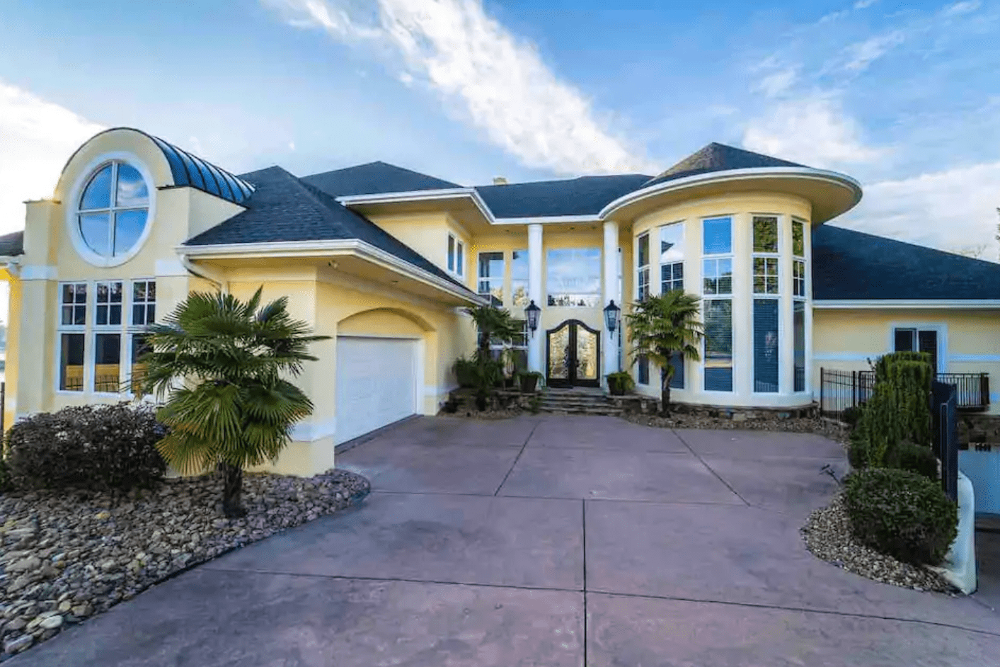 Large yellow house features arched windows and palm trees, with a wide driveway and clear blue sky above.