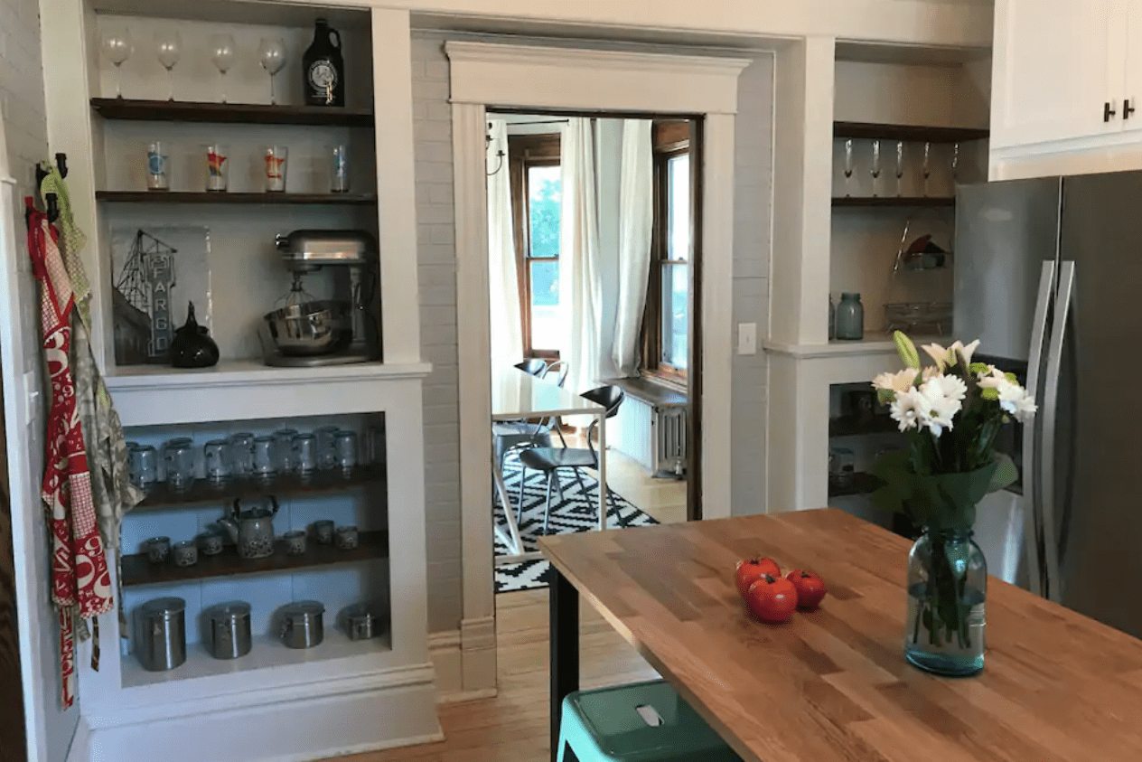 Kitchen shelves display glassware and appliances; counter holds tomatoes and flowers in a vase. Adjacent room has a table and chairs.