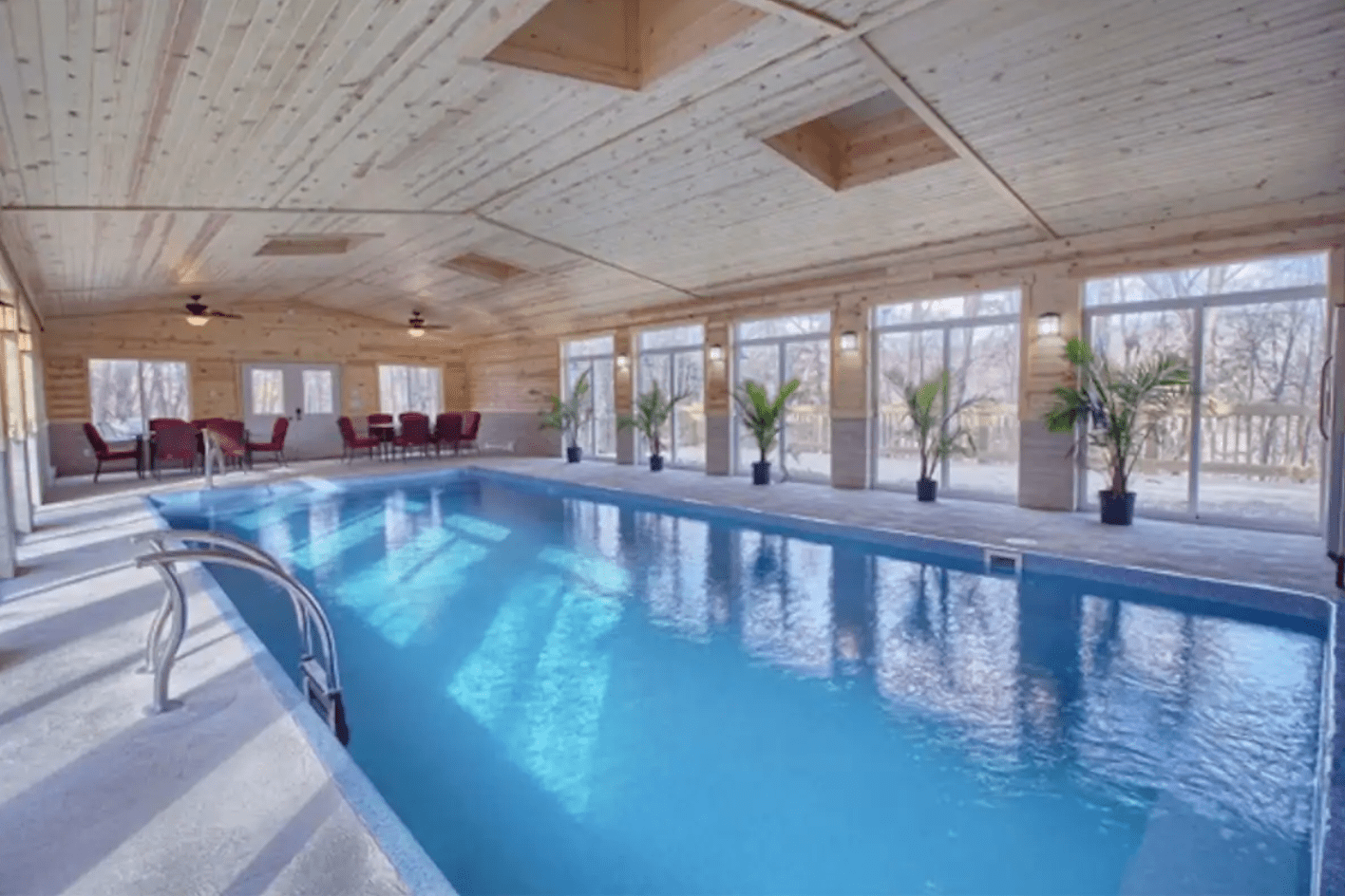 Indoor pool reflecting light, surrounded by chairs and tables under a wooden ceiling with large windows and potted plants lining the walls.