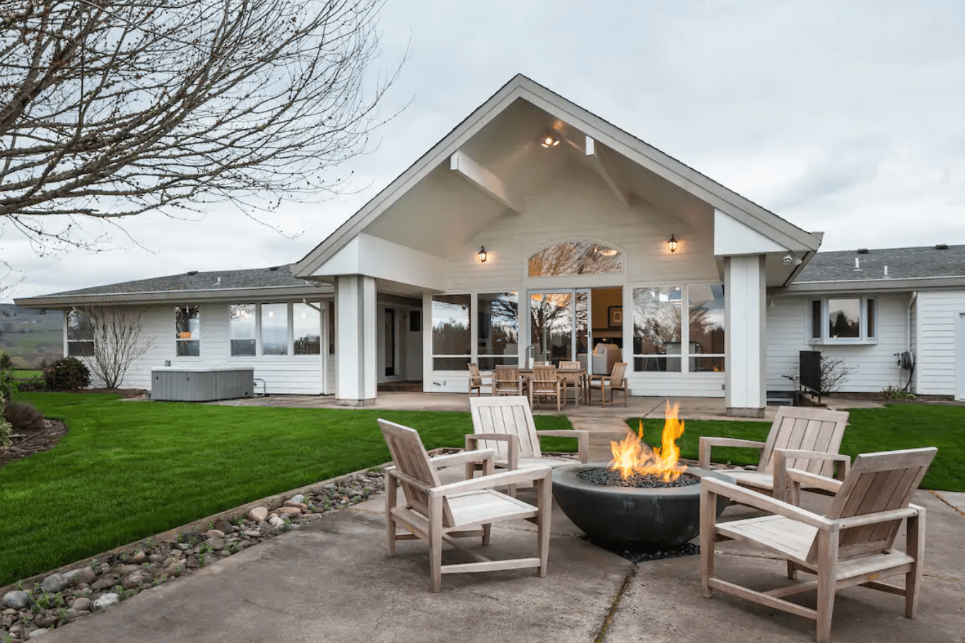 Fire pit burns surrounded by wooden chairs on concrete patio, in front of a white house with green lawn and cloudy skies.