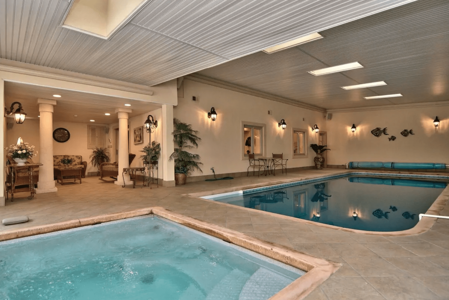 A spa bubbles adjacent to an indoor pool, surrounded by wicker furniture and decorative plants in a warmly lit room.