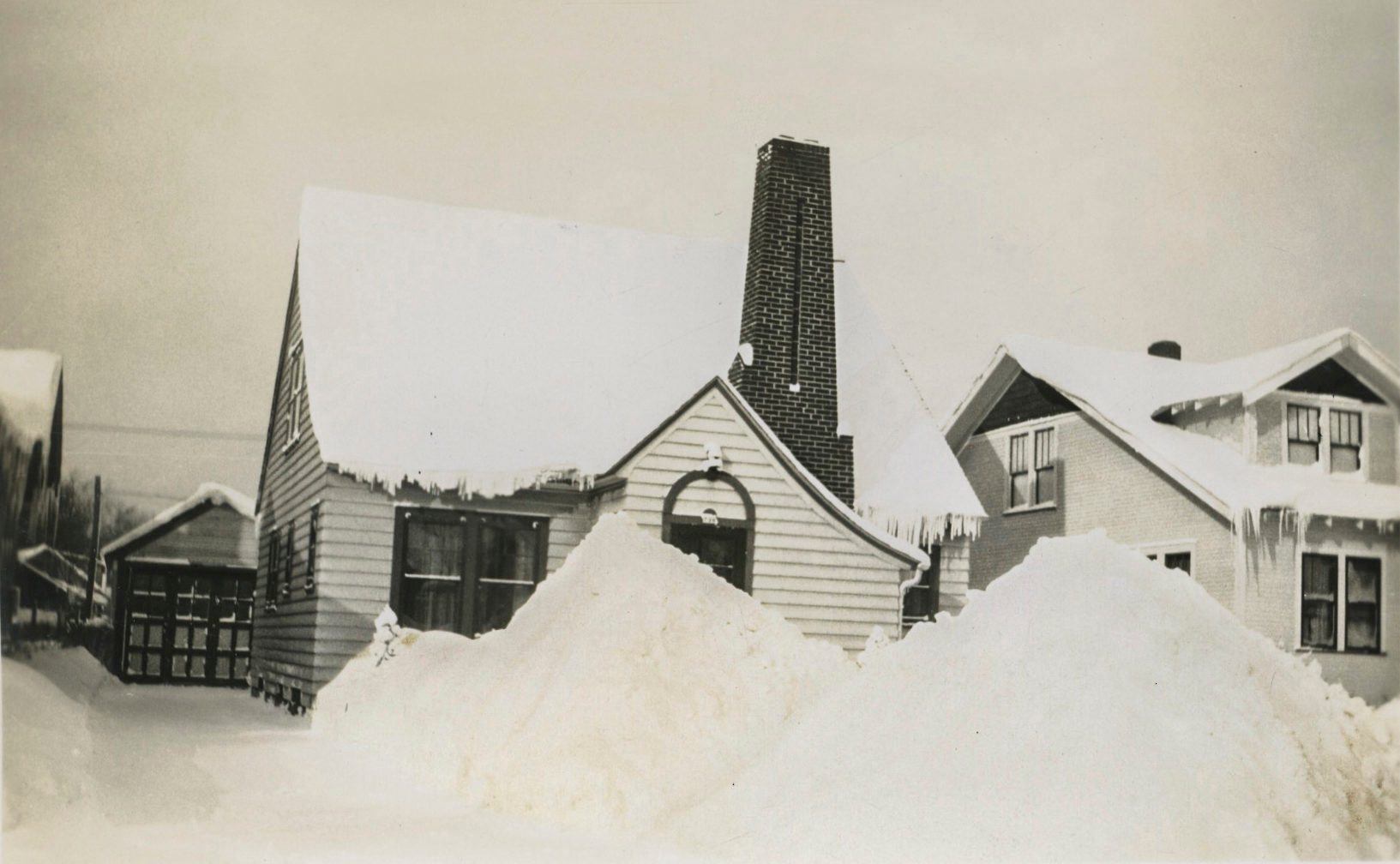 Houses covered in snow, surrounded by large snowdrifts, set in a wintry neighborhood with visible icicles hanging from roofs.